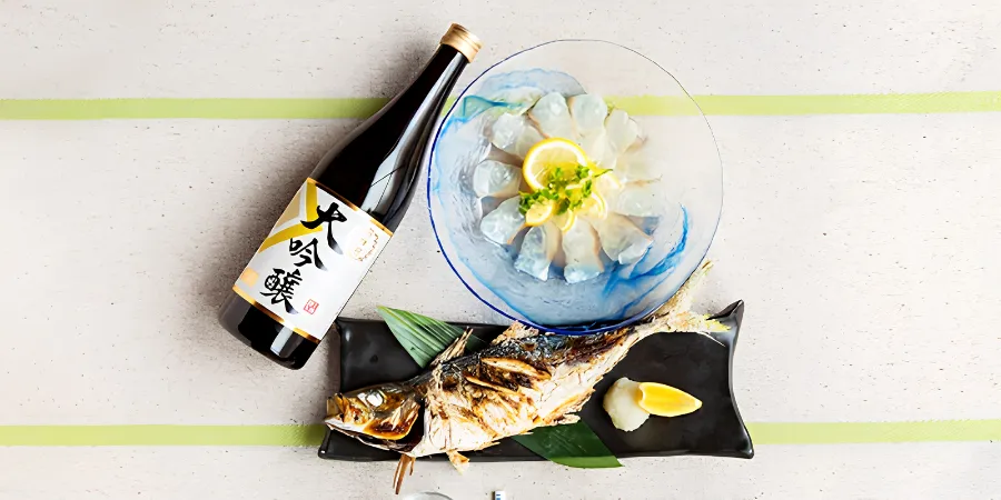 A bottle of Japanese sake beside a plate of thinly sliced sashimi garnished with lemon and herbs, with grilled fish, grated daikon, and lemon wedges arranged on a black serving plate.