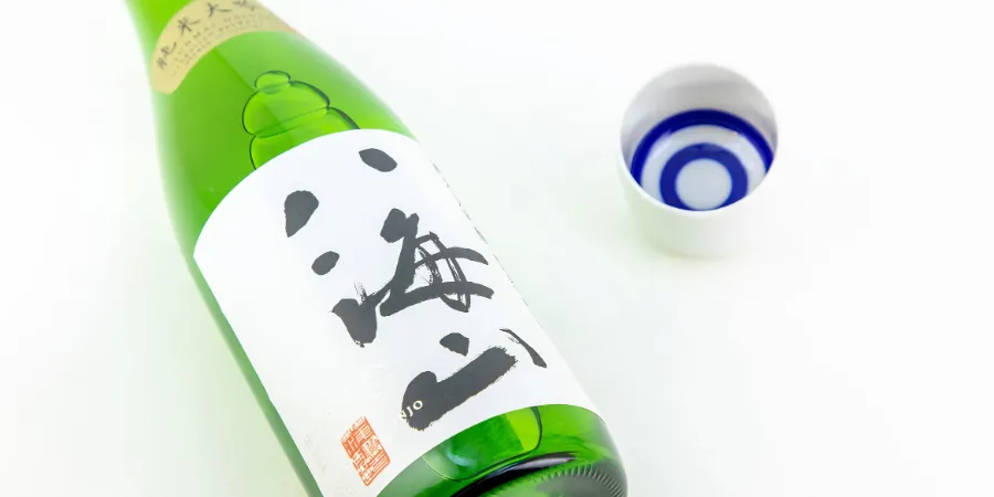 A bottle of Hakkaisan Japanese sake beside a blue-and-white sake cup on a white surface.