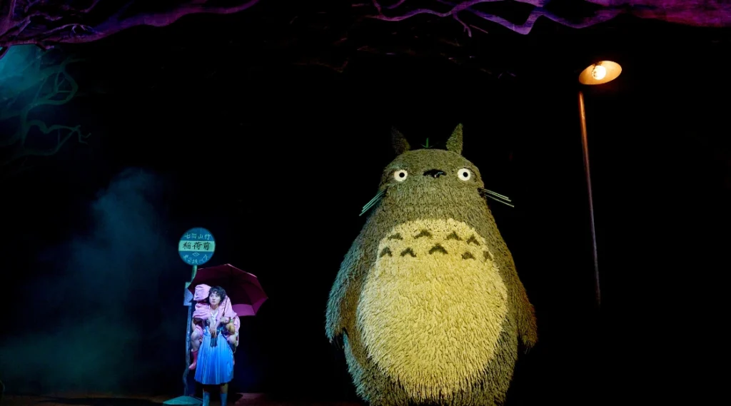 Helen Chong as Satsuki and Victoria Chen as Mei wait beside the giant Totoro puppet at a moonlit bus stop in My Neighbour Totoro at the Gillian Lynne Theatre.