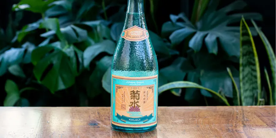 Kikusui Junmai Ginjo sake bottle displayed on a wooden table in front of lush tropical foliage.