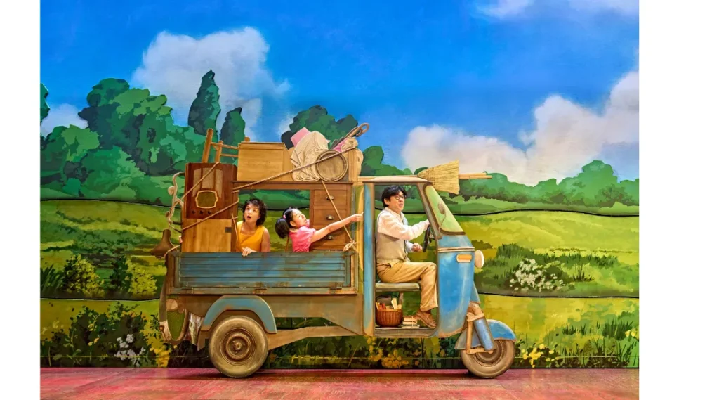 Victoria Chen as Mei, Ami Okumura Jones as Satsuki and Dai Tabuchi as Tatsuo ride in a blue moving truck through the countryside in My Neighbour Totoro at the Gillian Lynne Theatre.