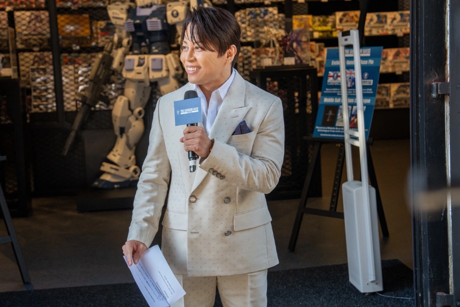 Mr Nishikawa at Gundam Store: Takanori Nishikawa speaking into a microphone at The Gundam Base London, with Gundam models and merchandise displayed behind him.