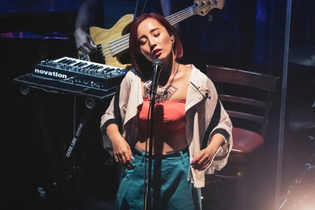 Nao Yoshioka performs on stage at Blue Note Tokyo, a microphone, with a bassist and keyboard equipment behind her.