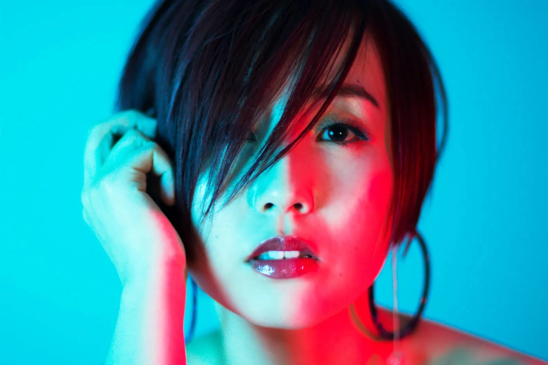 Nao Yoshioka in a close-up portrait, lit with vivid blue and red lighting, with short dark hair framing her face.