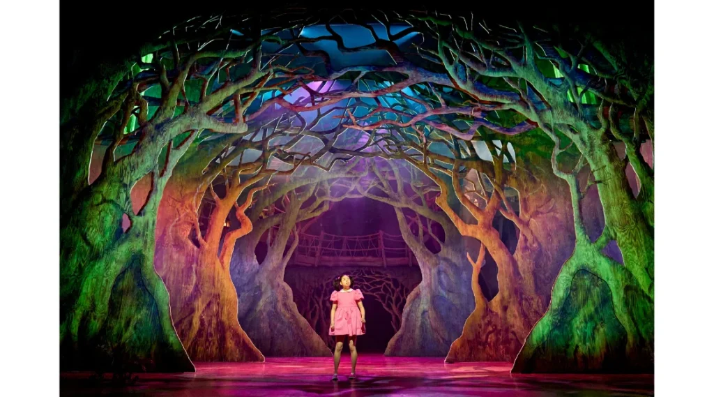Victoria Chen as Mei stands alone beneath a colourful canopy of towering trees in My Neighbour Totoro at the Gillian Lynne Theatre.