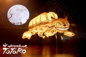 Illuminated Catbus puppet from My Neighbour Totoro on stage beneath a glowing full moon, with performers carrying it through a dark forest set.