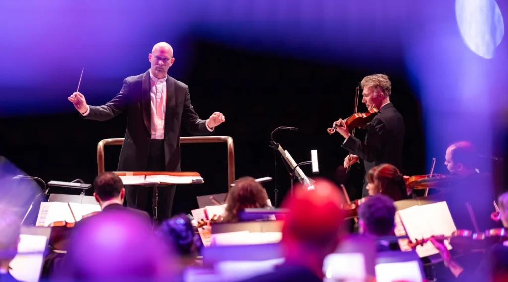 Michael England, Conductor leading The Sound of Epic Anime orchestra as a violin soloist performs at the front of the stage.