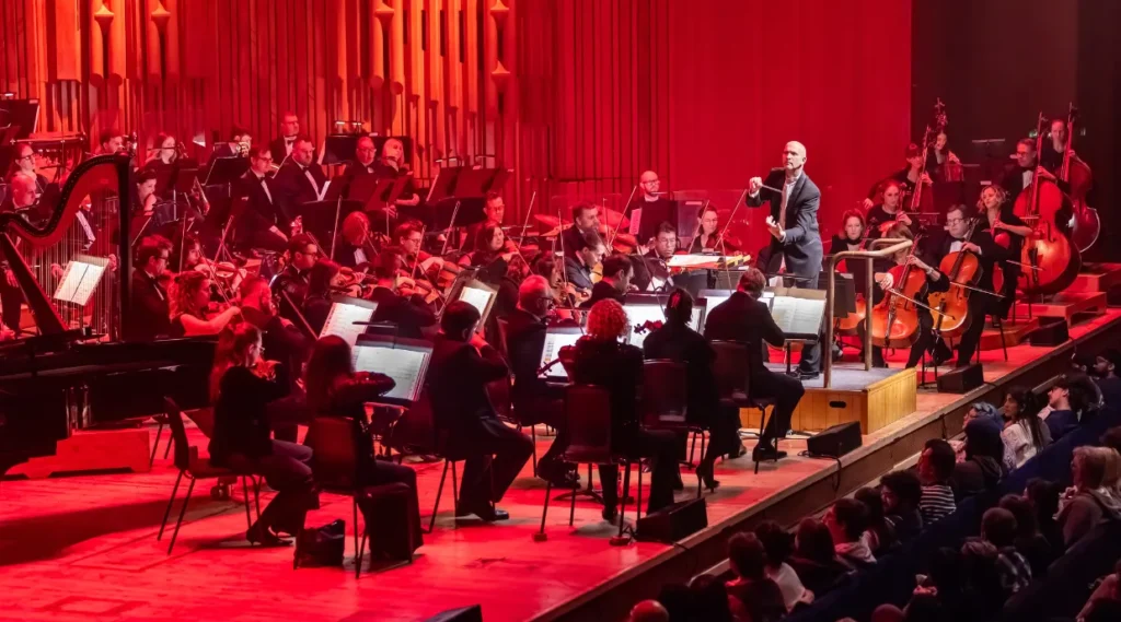 The Sound of Epic Anime full orchestra performing under dramatic red lighting as the conductor leads from the podium.