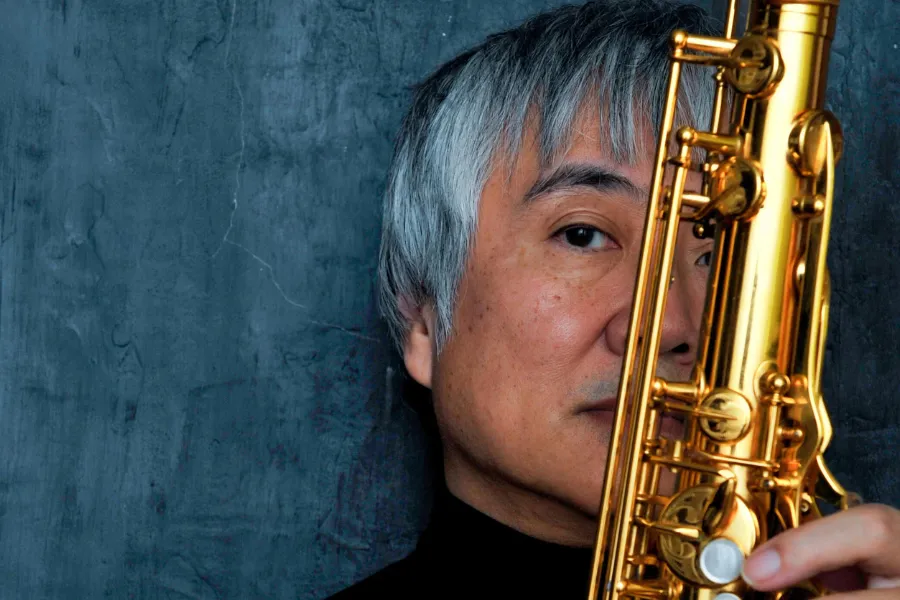 Yasuaki Shimizu playing a gold saxophone in a close-up portrait against a dark blue textured background