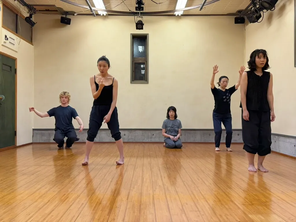 Workshop participants in dark clothes move slowly across a wooden studio floor, some crouching and some standing with arms raised
