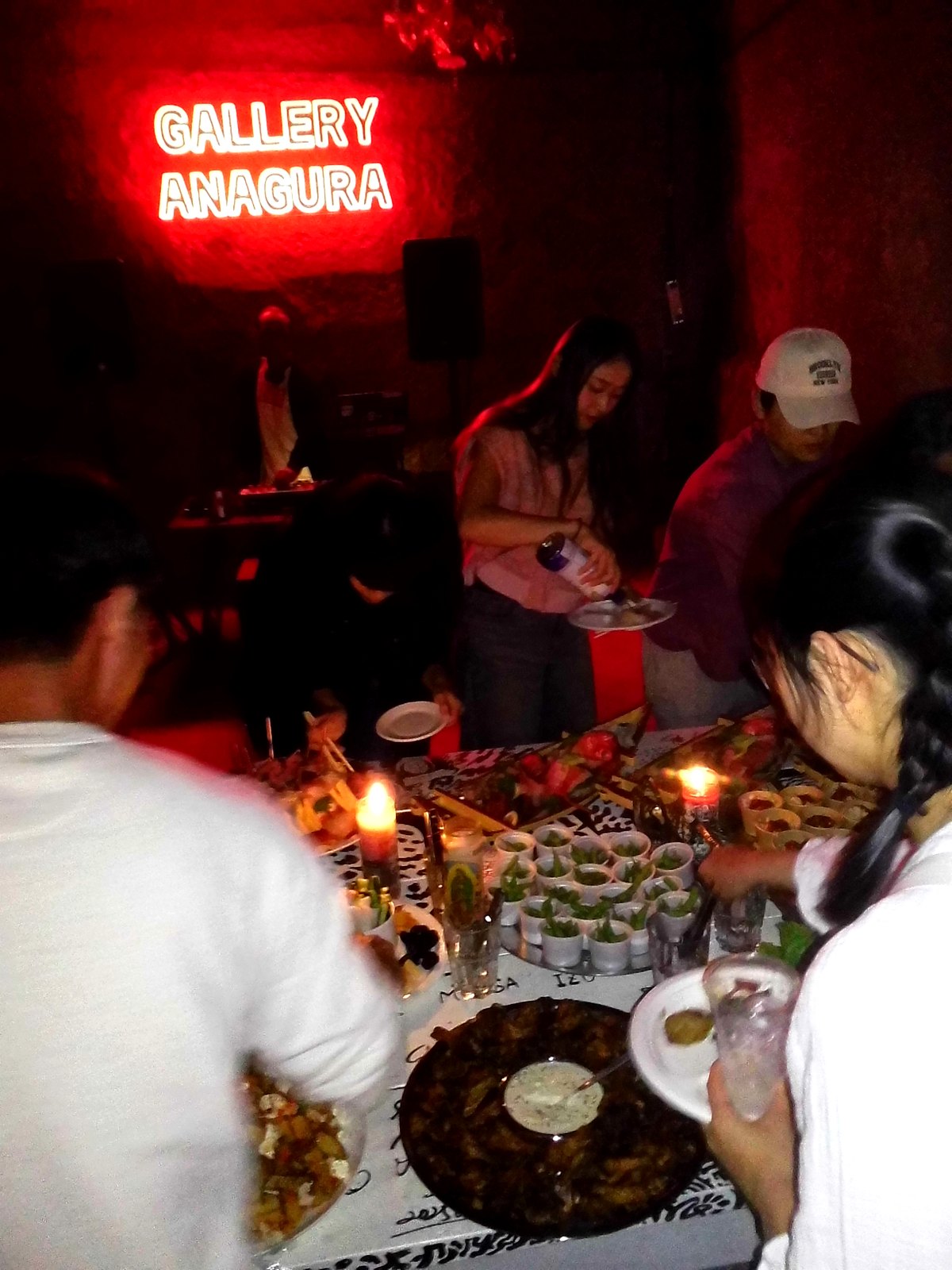 Long table of food lit by candles inside the Gallery Anagura cave bar in Shimoda, with a red neon sign on the rock wall and a DJ setup behind