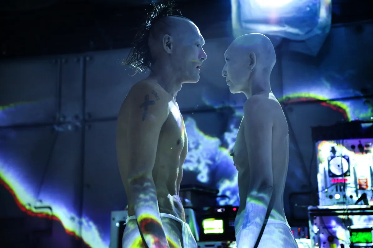 The two MUTSUMINEIRO dancers, heads shaved and bodies painted pale, face each other under blue and white projected light