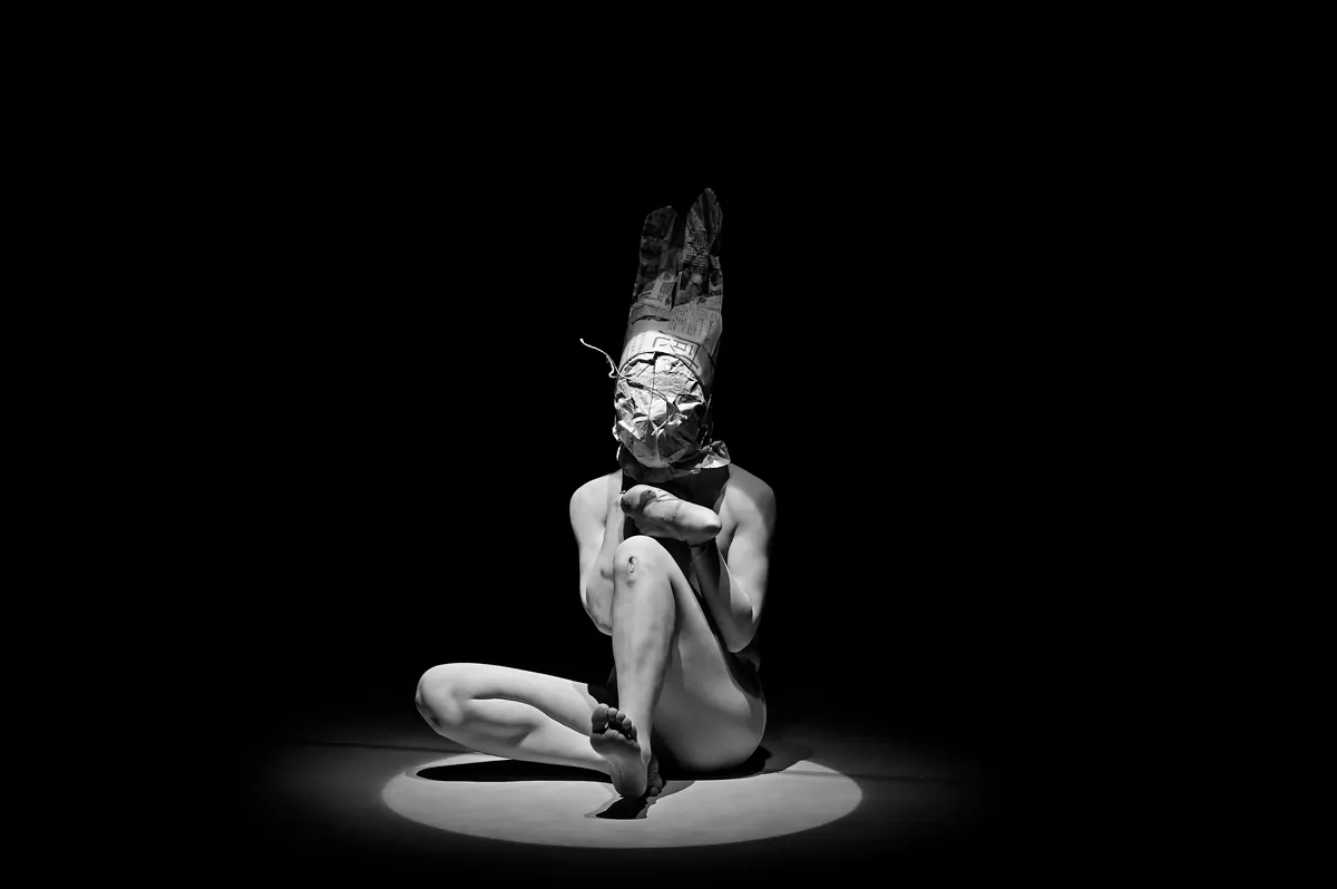 Makiko Takamatsu, wearing a tall rabbit eared mask, crouches in a single pool of white light on a black stage
