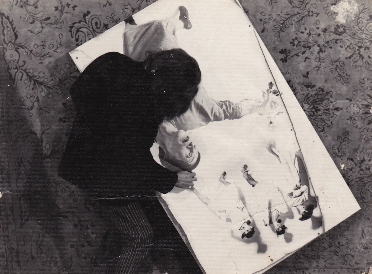 Seen from above, Kazuo Ohno in a dark coat lies across a large white board on a patterned floor, surrounded by scattered figures