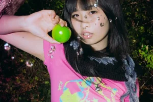 A model in a pink graphic T-shirt and fluffy black scarf holds up a green apple, from the Margins United 2026 merchandise shoot