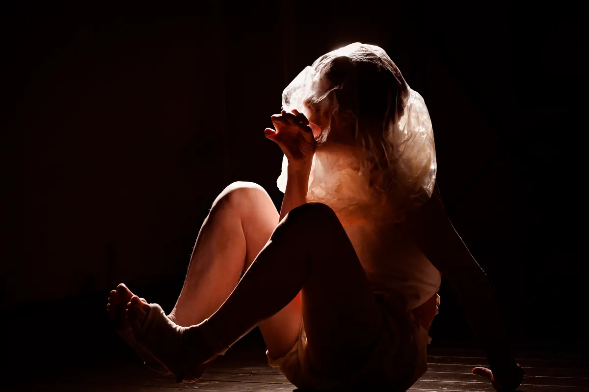 Mitsuyo Uesugi sits on a dark stage with her knees drawn up, a sheet of crumpled plastic over her head lit from behind