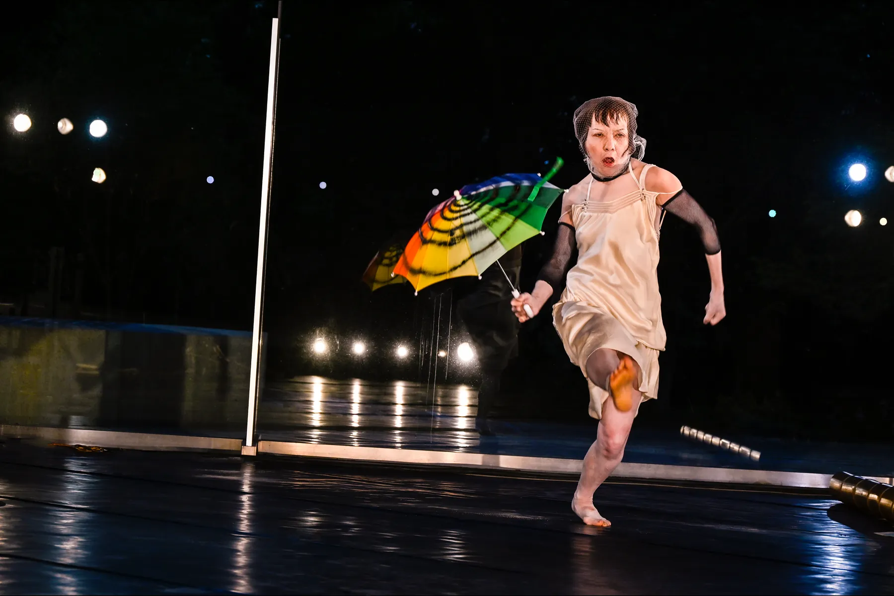 Mitsuyo Uesugi in a pale slip dress and hairnet runs barefoot across a wet outdoor stage at night, holding a rainbow striped umbrella