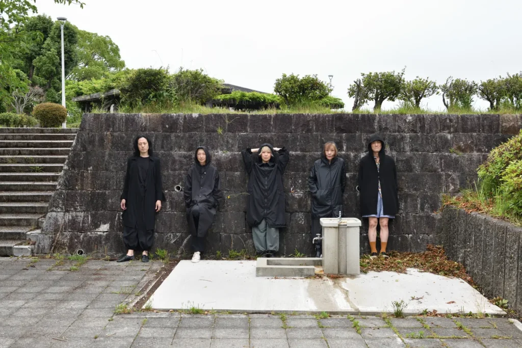 The five Mother Forest dancers in long black hooded coats lean against an old stone wall in a paved courtyard