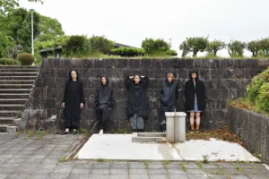 The five Mother Forest dancers in long black hooded coats lean against an old stone wall in a paved courtyard