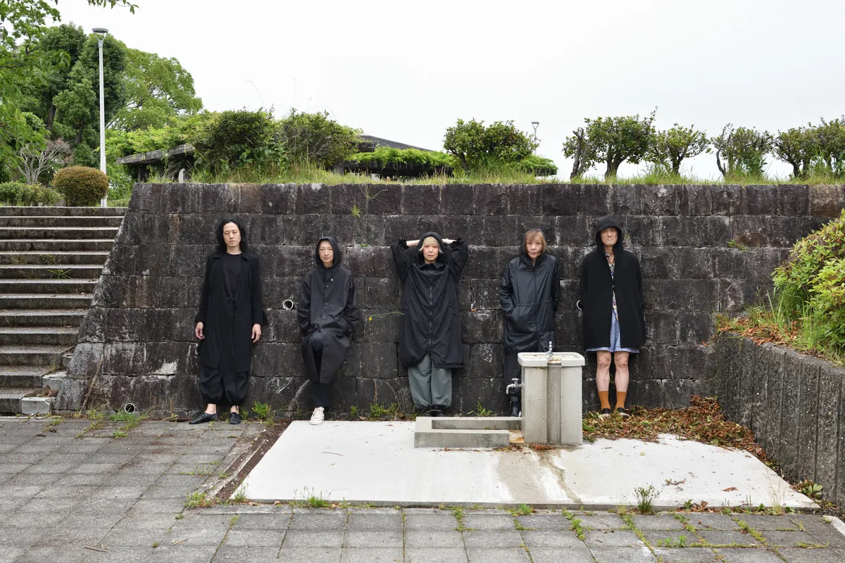 The five Mother Forest dancers in long black hooded coats lean against an old stone wall in a paved courtyard
