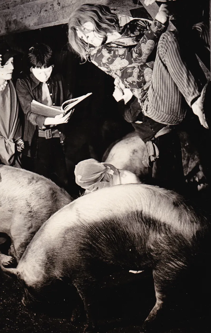 Dancers in a dark barn at the Yokodai Pig Farm, one leaping above the pigs while another reads from a book