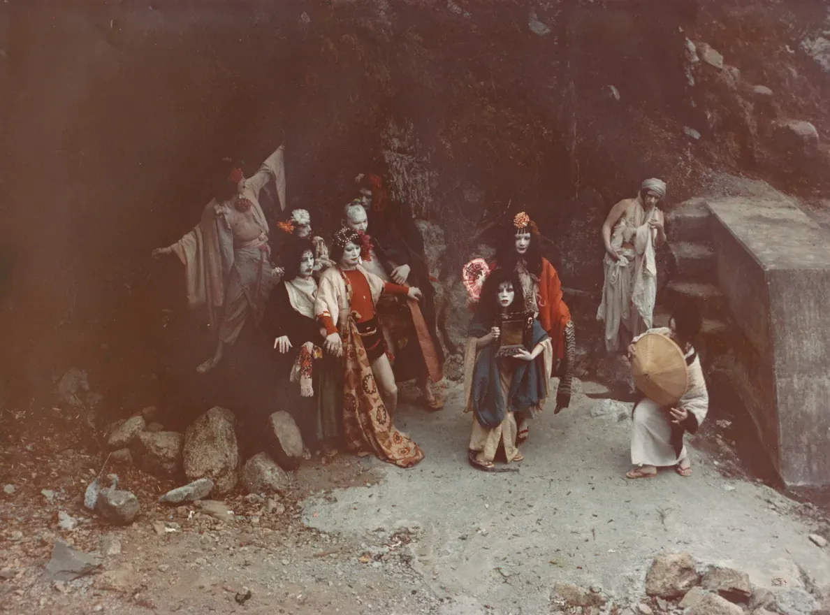 Kazuo Ohno’s students in colourful costumes and white face paint crowd a rocky mountain path in a colour still from the film