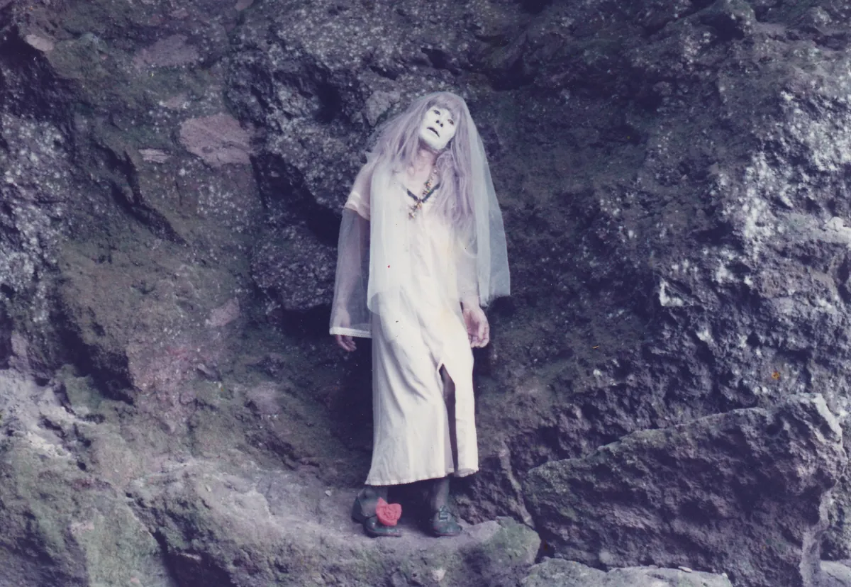 A figure in a long white dress, white veil and white face paint stands in the mouth of a dark rock cave