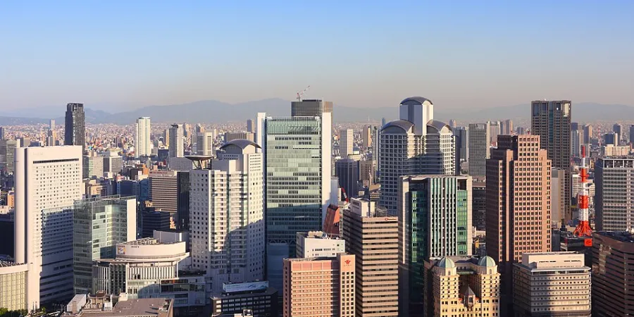 Panoramic view of the Osaka Umeda skyline, showing dense high-rise buildings and office towers with mountains in the background under a clear blue sky.