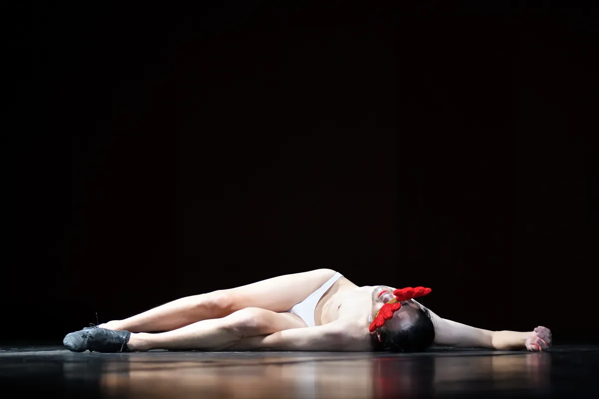 Seiji Tanaka lies stretched out on his back across a dark, glossy stage floor, a red headpiece over his face