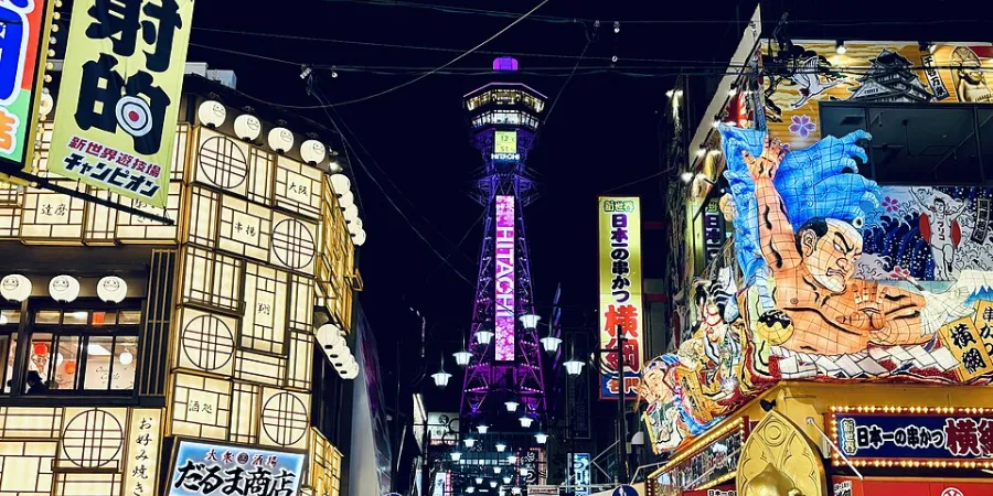 Nighttime view of Osaka's Shinsekai district with the illuminated Tsutenkaku Tower surrounded by colorful restaurants, Japanese signs and traditional entertainment venues.