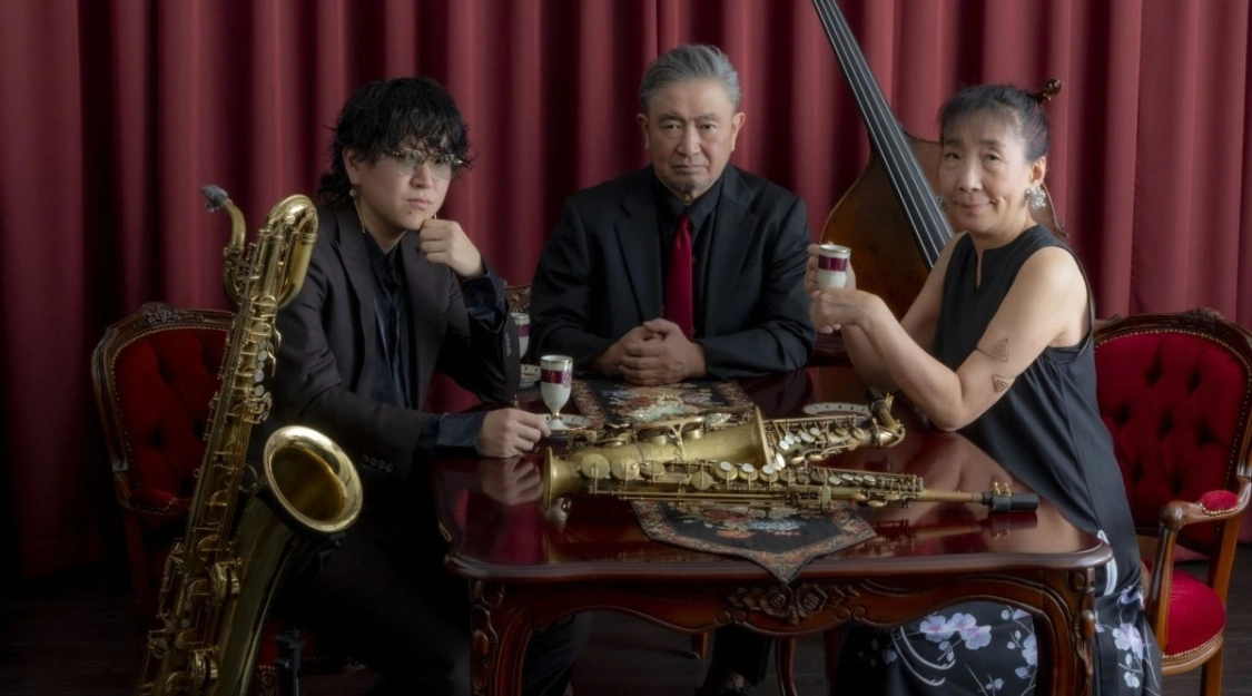 Saxophonist Sachi Hayasaka with Toshiki Nagata and Rio Nagata of the Tokyo family trio TReS, seated at a table with their saxophones and a double bass against red velvet curtains, ahead of their set at Haruki Murakami’s Jazz at Peter Cat at Barbican Hall on 21 November 2026