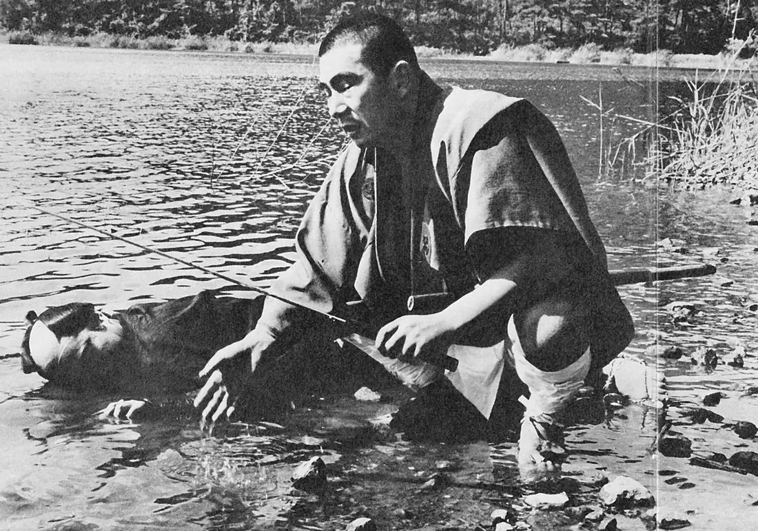 Shintaro Katsu as Ichi kneeling in shallow water beside a fallen opponent, a fishing rod lying across the frame, in The Tale of Zatoichi.