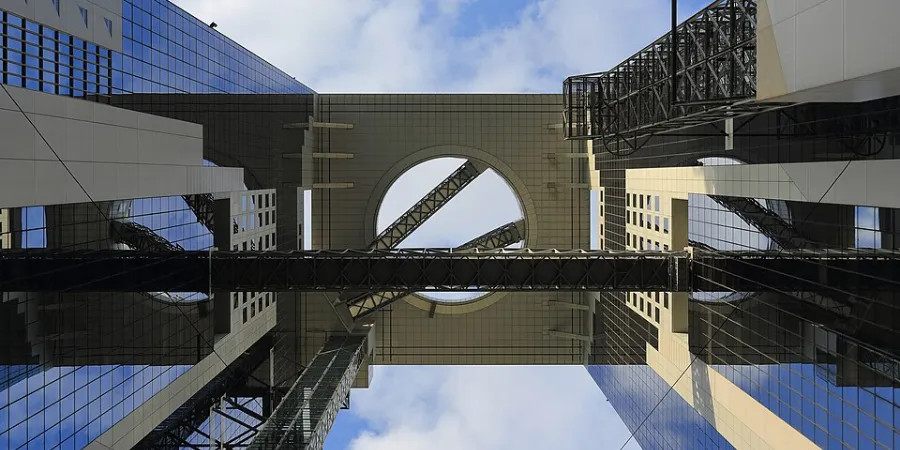 Looking upward through the geometric atriums of Osaka’s Umeda Sky Building, with mirrored glass towers, steel escalators and sky bridges framing a circular opening against a partly cloudy blue sky.