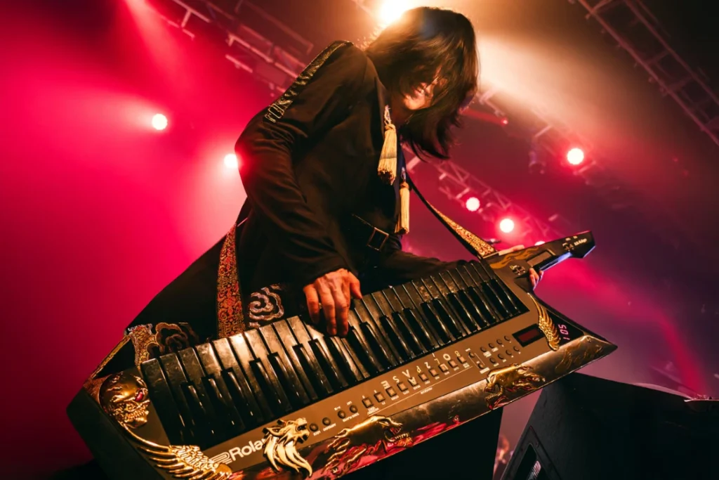 Naruto composer Yasuharu Takanashi leaning over his gold dragon Roland keytar under red stage lights