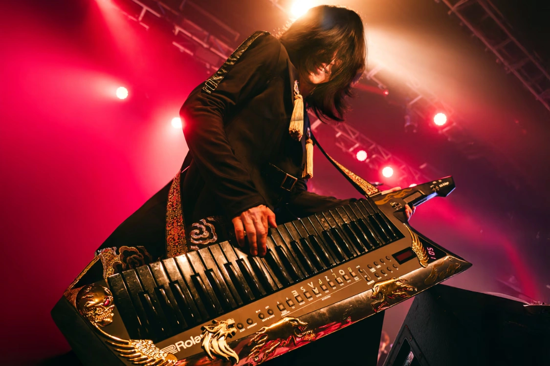 Naruto composer Yasuharu Takanashi leaning over his gold dragon Roland keytar under red stage lights