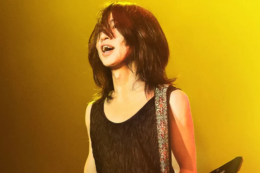 Yasuharu Takanashi singing on stage with Far East Groove, head tilted back under gold stage lighting, keytar strap over one shoulder