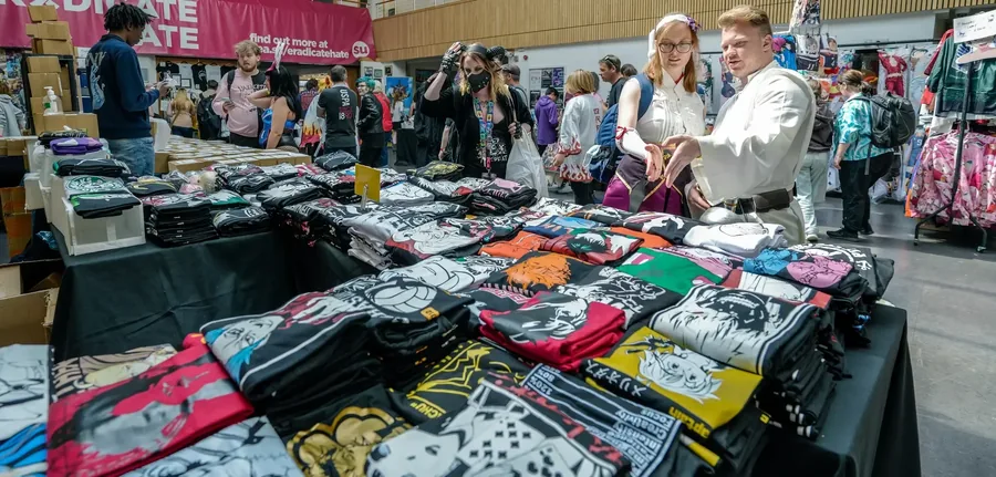 Anime T-shirts laid out on a trader's table at an anime convention, with cosplayers browsing the stalls behind