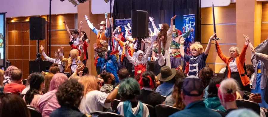 Dozens of cosplayers with their arms raised on stage at AnimeCon, watched by a seated crowd