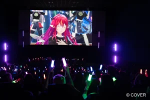 Wide view of a hololive cinema screening in Milan, a red haired VTuber on screen and the audience below with penlights raised