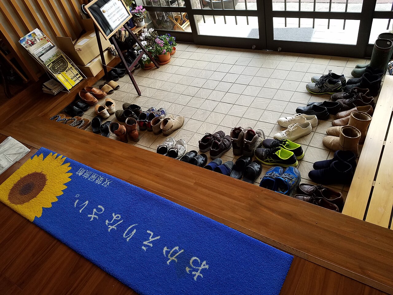 Guests’ shoes lined up in a Japanese inn entrance beside a mat reading okaerinasai, welcome home