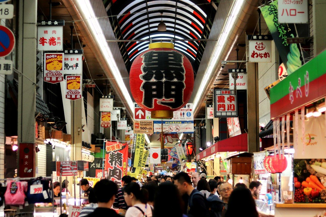 Shoppers under the covered arcade of Kuromon Ichiba Market in Osaka, with a large red lantern marked 黒門 hanging overhead