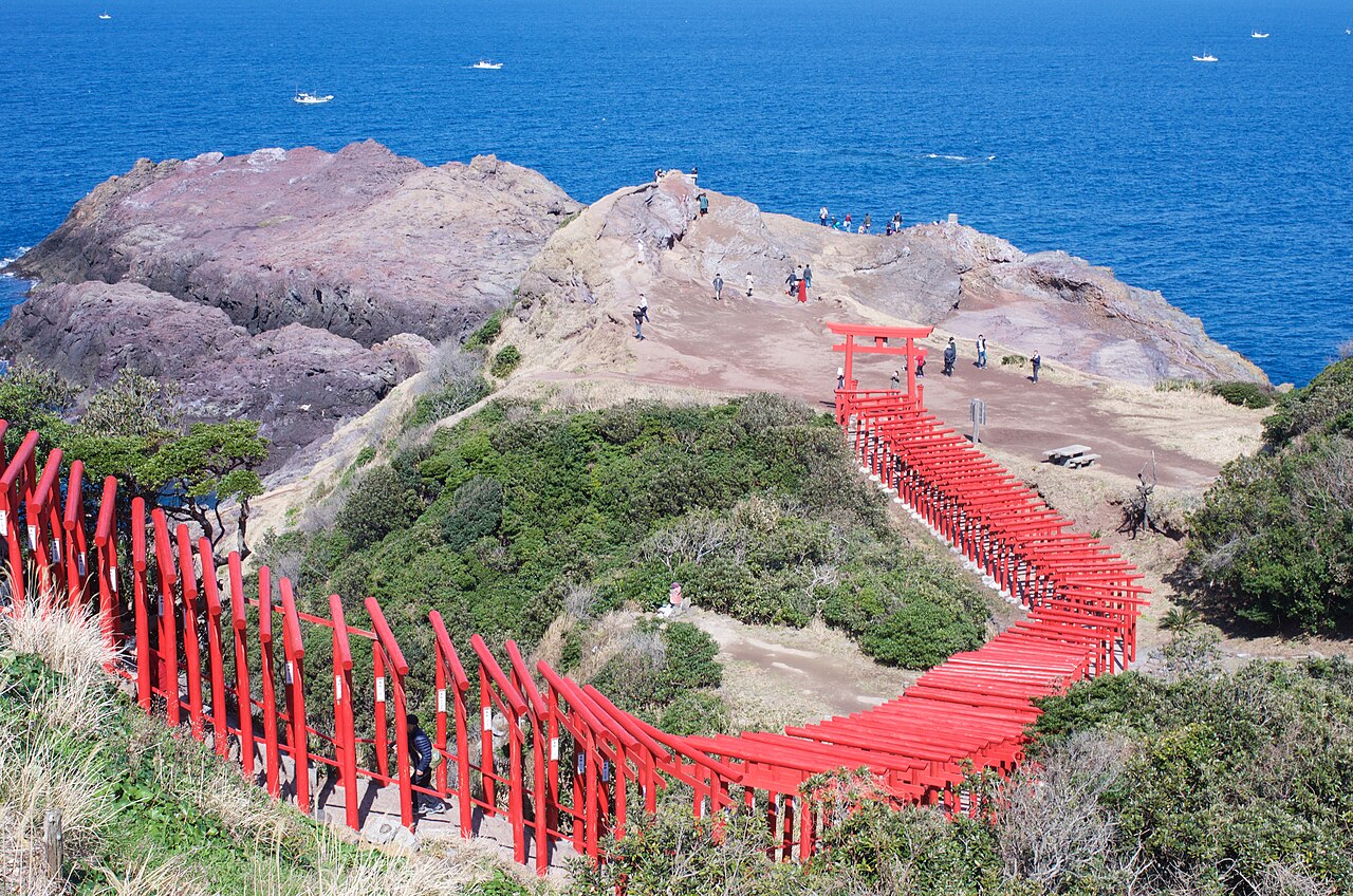 The red torii of Motonosumi Shrine curving down the hillside towards a rocky headland and the blue Sea of Japan