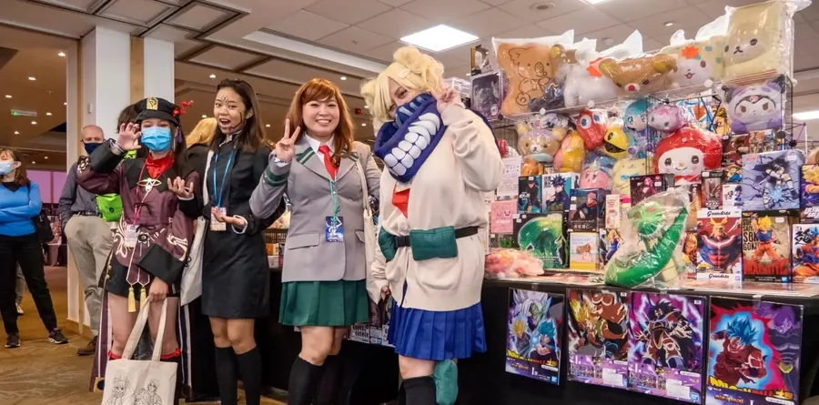Cosplayers dressed as Ochaco Uraraka and Himiko Toga from My Hero Academia and Hu Tao from Genshin Impact, posing beside a stall of anime figures and plush toys