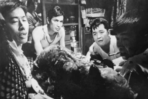 Black and white still from Shōhei Imamura's Pigs and Battleships, 1961, showing young men gathered round a table in a crowded Yokosuka bar