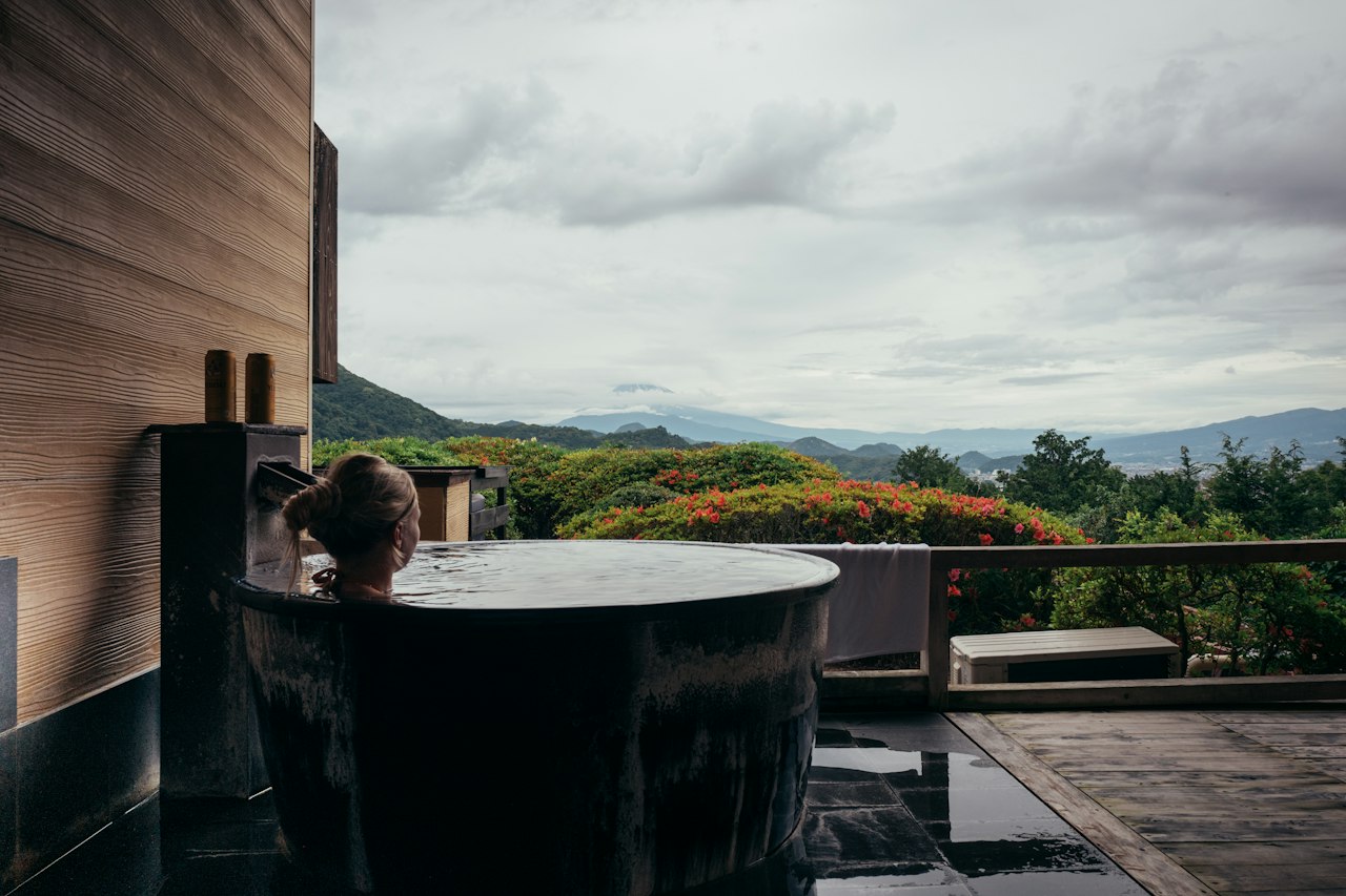 Guest soaking in a private outdoor hot spring tub on a wooden terrace looking out over forested mountains