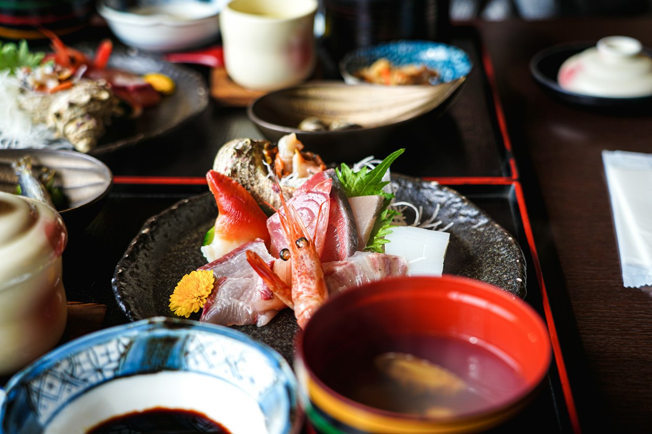 Sashimi of prawn, fish and shellfish on a dark ceramic plate surrounded by small dishes and lacquered trays for a multi course meal in Kyoto