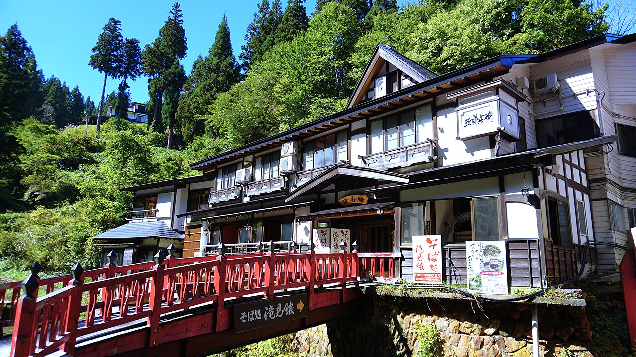 Multi storey wooden ryokan beside a red bridge over the river at Ginzan Onsen, surrounded by green forest