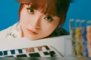 TC.KYLIE resting her chin on a synthesiser keyboard in front of a blue backdrop