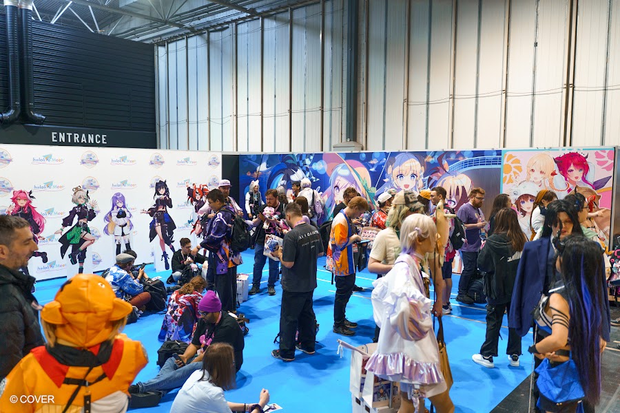 Visitors in cosplay and convention lanyards on the blue carpeted floor of the NEC hall near the entrance, beside the hololive Meet standee wall