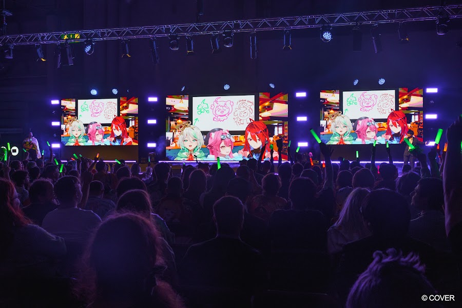 Three hololive English avatars on the Monarch Stage screens at VeXpo 2026, with the audience holding green penlights in front